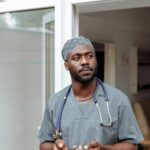 Portrait of a healthcare professional in scrubs holding a phone at a hospital entrance.
