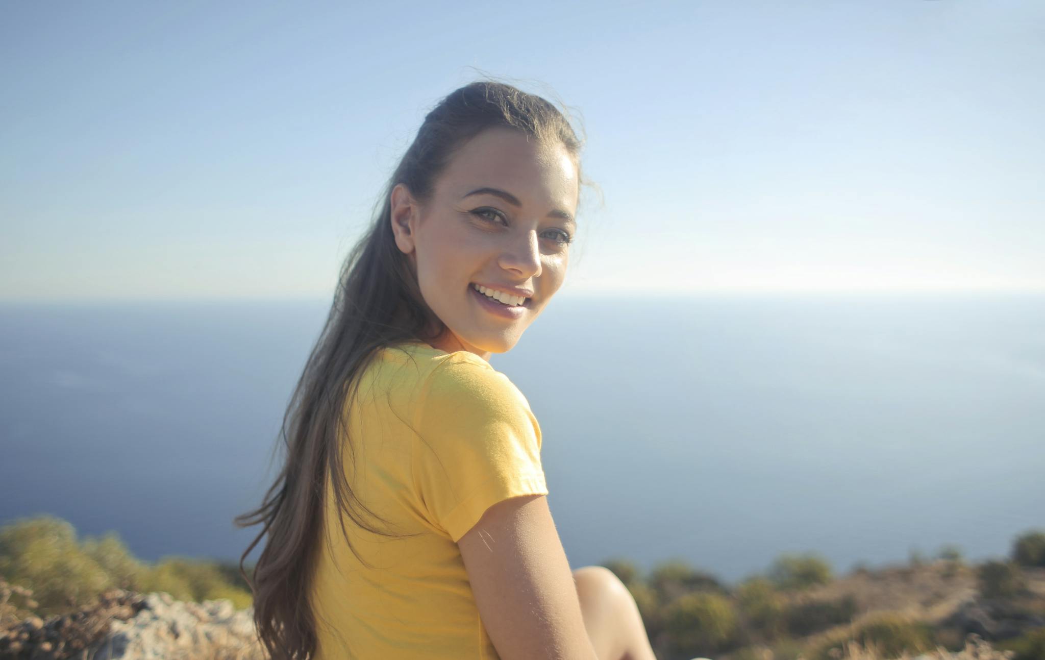 Young woman in a yellow shirt smiles by the sea, enjoying the outdoor view.
